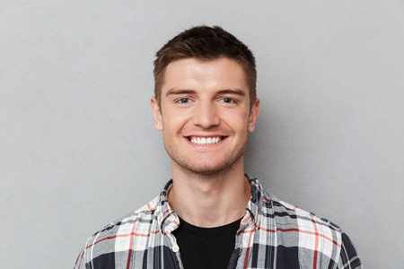 Close up portrait of a smiling young man in plaid shirt looking at camera over gray background