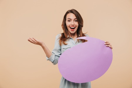 Image of happy young woman holding speech bubble posing isolated over beige background wall looking camera.の写真素材