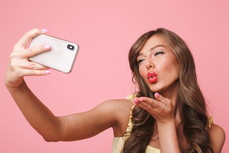 Photo of sexual pretty woman 20s with long brown hair smiling and giving air kiss at camera while taking selfie on mobile phone isolated over pink backgroundの写真素材