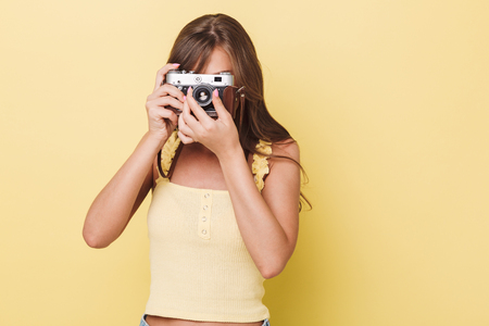 Portrait of a pretty young girl taking picture with a photo camera isolated over yellow backgroundの写真素材