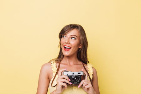 Portrait of a happy young girl holding photo camera and looking at copy space isolated over yellow backgroundの写真素材