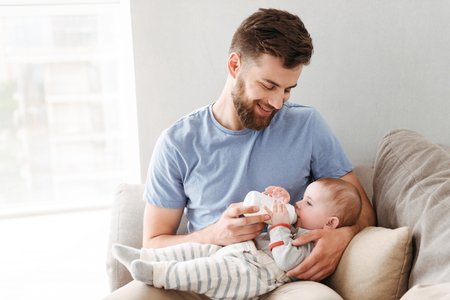 Image of young family. Father feeds her little child indoors at home.