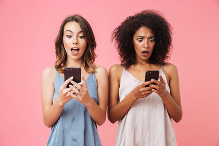Two surprised young girls using mobile phones while standing isolated over pink backgroundの写真素材