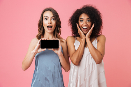 Image of pretty shocked young two women standing isolated over pink background showing display of mobile phone.の写真素材
