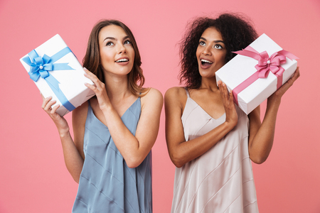 Image of happy young two women standing isolated over pink background holding gifts present boxes. Looking aside.の写真素材