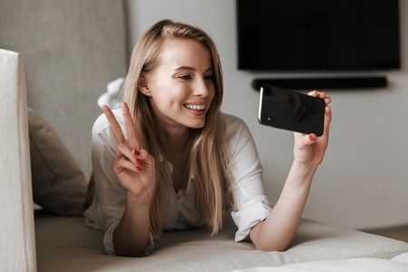 Image of happy cute young woman lies indoors at home talking by mobile phone make selfie.の写真素材