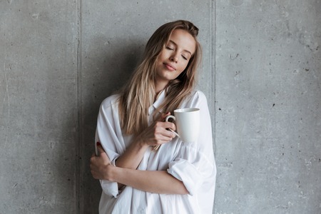 Picture of pleased young woman indoors over grey wall drinking coffee holding cup.の写真素材