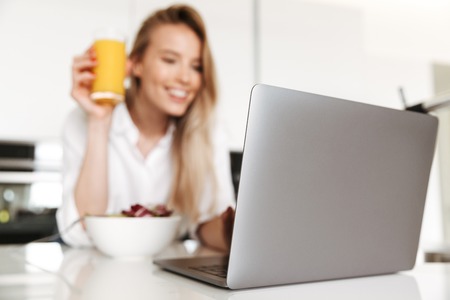 Close up of happy woman eating salad from a bowl and drinking orange juice while standing on a kitchen and watching movie on laptopの写真素材