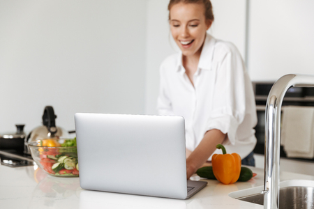 Cheerful young girl cooking salad while standing on a kitchen with laptop computerの写真素材