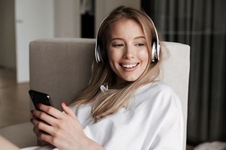 Smiling young woman in headphones using mobile phone while laying on a couch at homeの写真素材