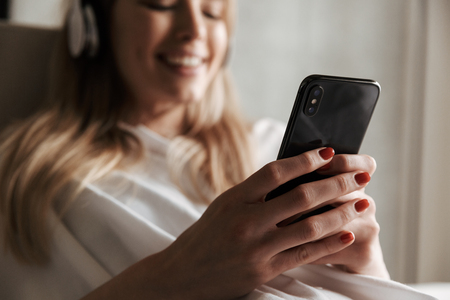 Close up of cheerful woman in headphones using mobile phone while laying on a couch at homeの写真素材