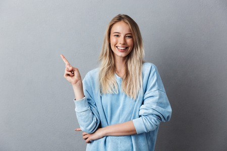 Portrait of a cheerful young girl in blue sweatshirt pointing at copy space isolated over gray backgroundの写真素材