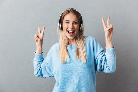 Portrait of a cheerful young blonde girl listening to music with headphones and showing peace gesture isolated over gray backgroundの写真素材