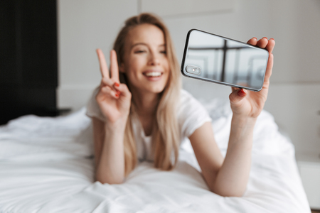 Photo of cute beautiful young woman indoors at home make selfie by mobile phone showing peace gesture.の写真素材