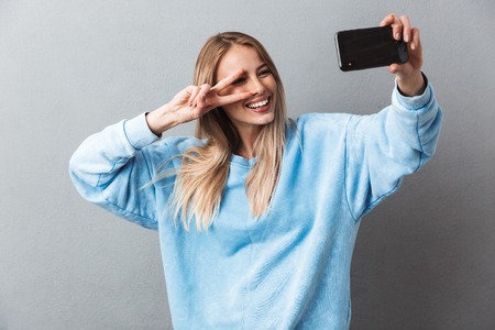 Smiling young blonde girl taking a selfie and showing peace gesture isolated over gray background