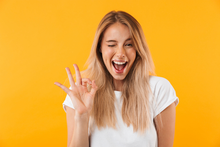 Portrait of a happy young blonde girl showing ok gesture and winking isolated over yellow backgroundの写真素材