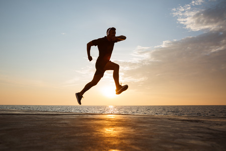 Silhouette of active sporty man 30s jumping and running along pier at seaside and listening to music via wireless headphones during sunriseの写真素材