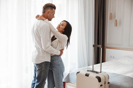 Image of multiethnic couple man and woman wearing casual clothing hugging together in hotel room with big luggage. Vacation conceptの写真素材