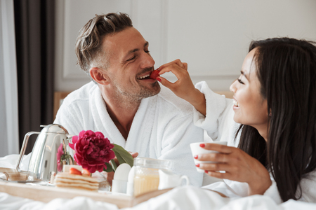 Image of beautiful asian woman feeding caucasian man with strawberry while lying happily in bed at hotel apartment wearing white bathrobeの写真素材