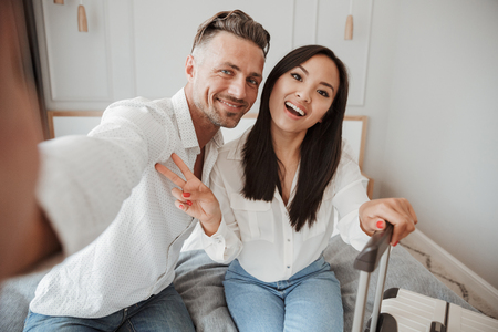 Excited young couple taking selfie with mobile phone while sitting together on bed at a hotel room with a suitcaseの写真素材