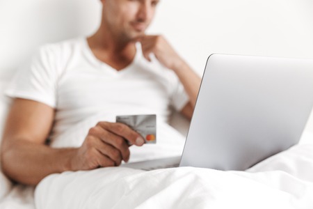 Close up of a concentrated man using laptop computer while sitting in bed and holding credit cardの写真素材