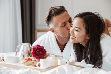 Lovely young couple dressed in bathrobes having romantic breakfast while lying on bedの写真素材