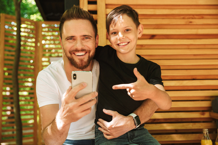 Image of happy young father sitting with his little son outdoors in cafe nature using mobile phone pointing.の写真素材