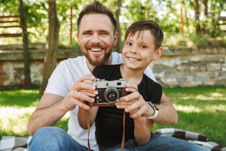 Photo of young father sitting with his little son outdoors in park nature holding camera photographing.の写真素材