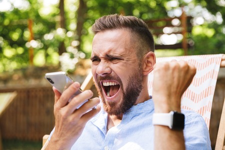 Photo of happy young bearded man outdoors using mobile phone make winner gesture.の写真素材