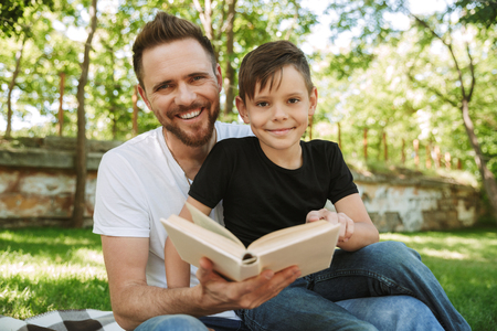 Image of happy young father sitting with his little son outdoors in park nature reading book.の写真素材