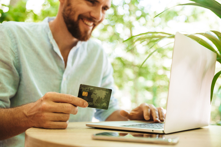 Photo of smiling emotional young bearded man outdoors using laptop computer holding credit card.の写真素材