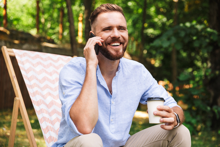Photo of happy young bearded man outdoors talking by mobile phone drinking coffee.の写真素材
