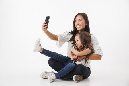 Image of emotional happy young asian woman mother with her little girl child daughter sitting isolated over white wall background make selfie by mobile phone.の写真素材