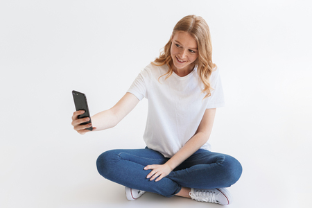 Picture of pretty young cute redhead woman sitting isolated over white wall background. Looking aside make selfie by mobile phone.の写真素材