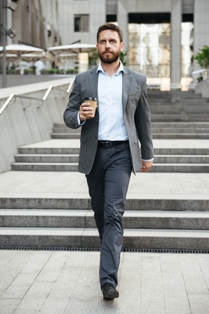 Photo of successful businessman 40s in gray suit walking down stairs of modern business center, with takeaway coffee in handの写真素材