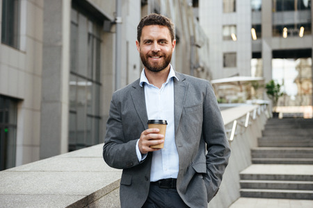Photo of successful businessman 40s in gray suit looking at you while standing and drinking takeaway coffee in front of modern business centerの写真素材
