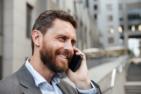 Portrait closeup of smiling businesslike man 40s in gray suit speaking on mobile phone while standing near office building on backgroundの写真素材