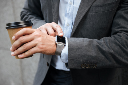 Cropped photo of european businessman in gray suit and white shirt standing against granite wall and touching smartwatch while drinking takeaway coffeeの写真素材