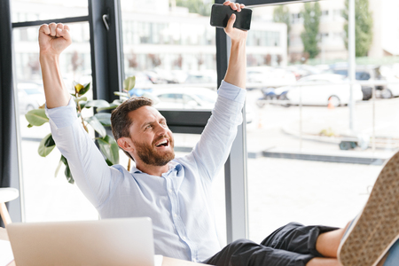 Image of happy cheerful bearded man in office working looking aside holding mobile phone.の写真素材