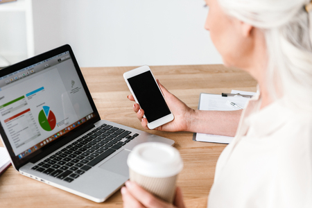 Close up of confident mature business woman working on laptop computer and drinking coffee while sitting at the desk in office and holding mobile phoneの写真素材