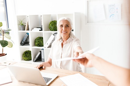 Smiling mature busienss woman giving documents while sitting at the desk with laptop computer in officeの写真素材