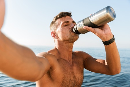 Tired handsome shirtless sportsman taking a selfie while standing at the beach and drinking water...
