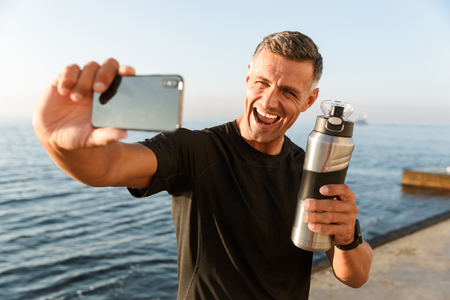 Image of handsome mature sportsman take a selfie with bottle of water on the beach.の写真素材
