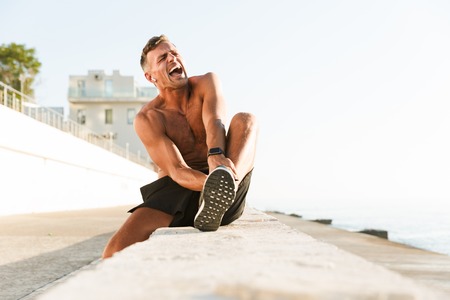 Image of sportsman on the beach outdoors listening music with earphones touching his leg because of...