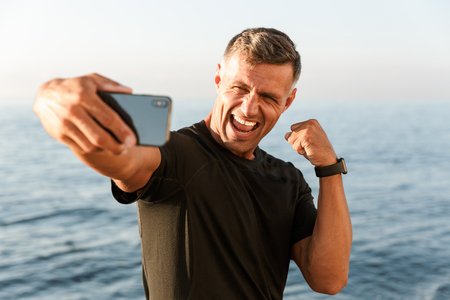 Cheerful handsome shirtless sportsman taking a selfie while standing at the beach and flexing biceps Cheerful handsome shirtless sportsman taking a selfie while standing at the beach and flexing biceps