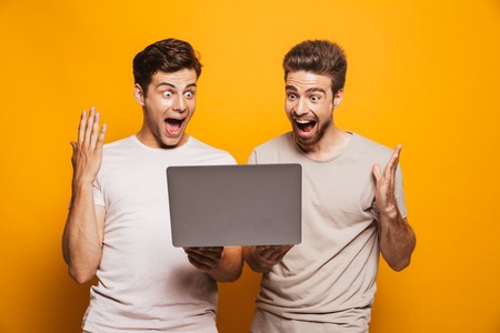 Portrait of a two cheerful young men best friends looking at laptop computer and celebrating isolated over yellow backgroundの写真素材