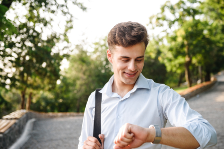 Smiling young man in shirt walking at the city park and looking at his wristwatchの写真素材