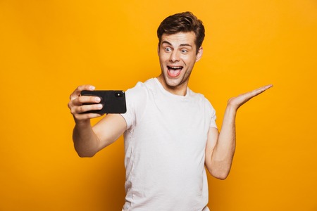 Portrait of a cheerful young man taking a selfie isolated over yellow background, celebrating successの写真素材