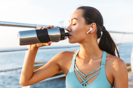 Close up of pretty asian sportswoman in earphones sitting outdoors at the beach and drinking from a water bottleの写真素材