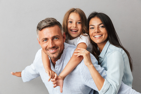 Portrait of a happy family father, mother, little daughter having fun together, father holding little daughter on his back isolated over gray backgroundの写真素材
